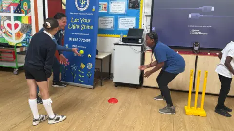 Sonny Baker plays catch with three school students in a large classroom.