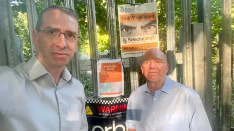 Hertfordshire Liberal Democrats Two people stand in front of a metal security fence displaying several notices, including a Network Rail "Footbridge Closed" sign and a warning poster about theft. The photograph was taken at the entrance to the closed Oxhey Grange footbridge, with trees and foliage visible behind the fence.