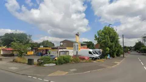 Street-corner view of a suburban road junction on a bright day, with a small commercial building and forecourt on the left, several parked vehicles including white vans and cars, trees and shrubs along the roadside, and a wide blue sky filled with large white clouds. A two-lane road extends into the distance past residential and commercial properties.