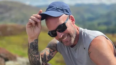 A man smiles as he lifts his cap to reveal his bald head. He is wearing a grey tank top and sunglasses and has a lot of tattoos on his arms. The countryside is blurred in the background.