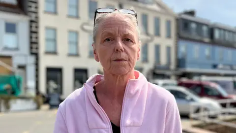 Tracy Benn is looking seriously into the camera. She is wearing a pink fleece and has glasses on her head. She is standing behind Gorey pier.