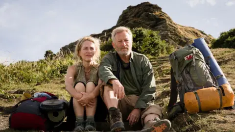 Gillian Anderson and Jason Issacs sit on a rocky outcrop with their backpacks looking at the view from the South West Coast Path.