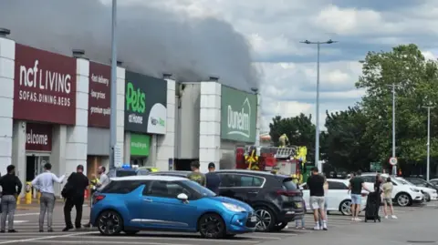 Crowds of shoppers look on as emergency service workers attend a fire at a store in a retail park. Smoke can be seen billowing from the Dunelm store. 