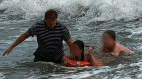 Jo Drake A man in a black and white striped shirt helping two young people in the water. One of the young people is in an orange hoop.