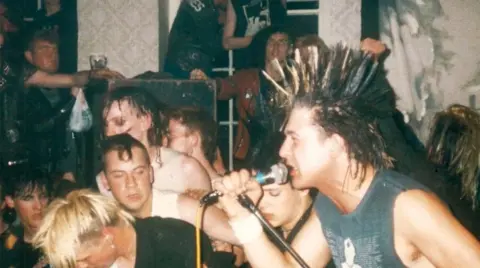 Historic England/Home of Metal A man with a mohawk, who is a member of the band Conflict, sings enthusiastically into a microphone, surrounded by people in the pub