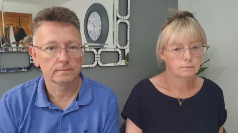 Mr and Mrs Glasser sitting beside each other. He has short grey hair and clear glasses. He is wearing a blue polo shirt. Beside him is his wife who has her blonde hair tied back and has a blonde fringe. She is wearing a black round neck t-shirt and gold jewellery. She has gold metal glasses. Behind them is a grey wall with a mirror on it.