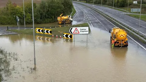 Councilllor Jon Abbott Yellow tank vehicles pump water out of a flooded road junction with a sign saying Oakley Clapham