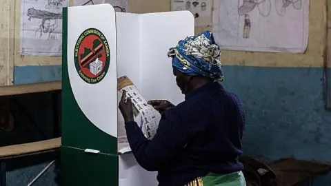 A person stands inside a polling booth in a classroom-like setting, marking a ballot. The room has worn walls, wooden desks, and hand‑drawn posters displayed on the walls. 