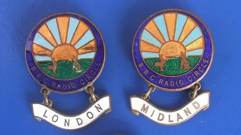 Stuart Williams/Flickr Two badges showing a drawing of a semi-circular sun with rays coming out of it. Two brown rabbits are sitting in front, looking at the sun while sitting on grass. There is a blue band around the picture, with the words BBC RADIO CIRCLE iin gold lettering. There is a white name plaque beneath - one has the word LONDON in black writing while the other has MIDLAND.