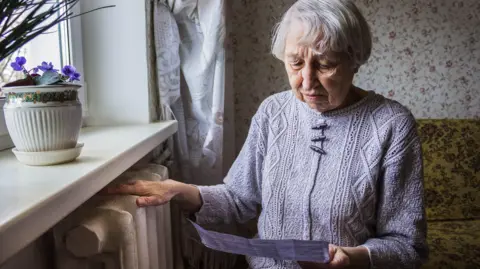 An elderly lady in her front room looks anxiously at a paper bill in her hand, with her other hand resting on a radiator. She has silver hair and is wearing a knitted jumper. 