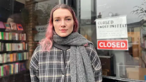 A woman with long pink hair and a grey striped jacket stands outside a shop. Behind the glass are rows of books. A sign on the door reads 'Closed Indefinitely'