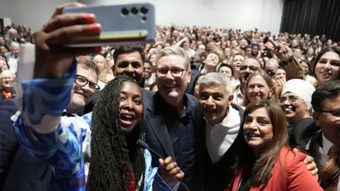 PA Media Prime Minister Sir Keir Starmer poses for a selfie with Dawn Butler, MP for Brent East and Mayor of London Sadiq Khan at a reception during the Labour Party Conference in Liverpool. A huge crowd of people stand behind the Labour politicians