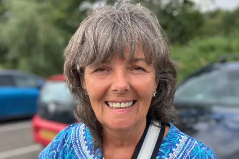 A woman with shoulder-length hair smiling for the camera in a car park.
