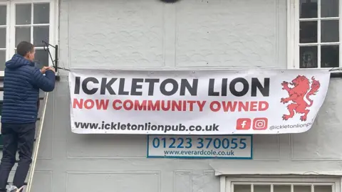 A man is standing on a ladder, against the Ickleton Lion pub, while hanging up a sign. It reads 'Ickleton Lion, now community owned'. 