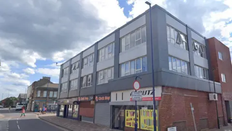 Google How the current building looks. It is a three-storey building with shops on the ground floor. It is brick built but the the front and side of the upper two floors are clad in contrasting light and dark grey panels. It has a flat roof.