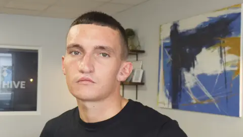 Image of a young man in a black t-shirt, staring directly into the camera. He has short cropped black hair and is clean shaven. He is stood in a room with a large abstract blue, white and black painting behind him, and a shelf with smaller objects on it. 