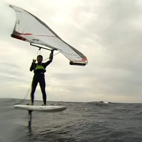 Martin Gaveriaux A wingfoiler stands balanced on a hydrofoil board above choppy sea, holding an inflated wing overhead while riding across the water under a cloudy sky.