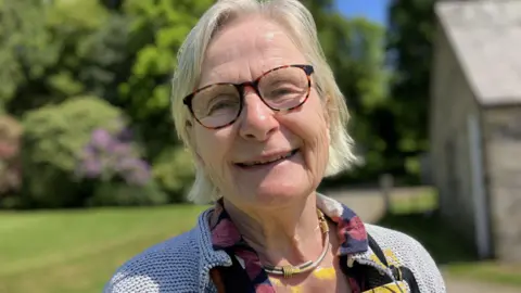 A woman with blonde/white hair, wearing glasses and smiling at the camera. She is wearing a choker style necklace and a flowery shirt with a cardigan over it. 