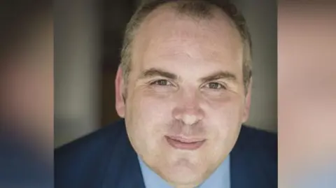 Paul Rosser Man looking directly at the camera. He is wearing a yellow tie with a light blue shirt and a dark blue suit.