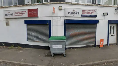 Google The business is pictured during the day with metal shutters pulled down. The building is white and with blue paint above the shutters with a white sign.