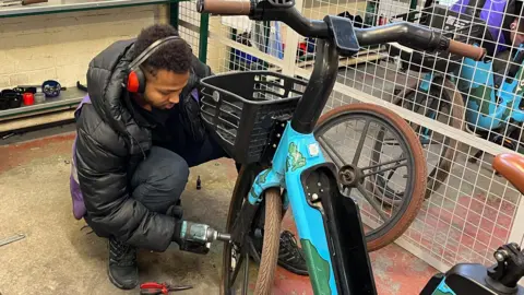 A worker in ear protectors is crouched infront of a blue bike and is tightening the wheel