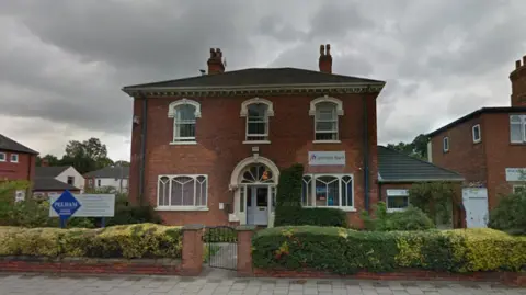 A double-fronted red-brick building with five large windows stands behind a small brick wall and yellow and green privet hedge. A blue and white sign is on the left of the image.