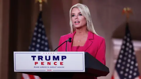 Pam Bondi speaks at the Republican National Convention in 2020. She is standing in front of a lectern with the words "Trump-Pence" on it. She has straight blonde hair and is wearing a pink blazer.