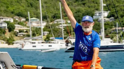 PA Media Frank Rothwell with a grey beard after arriving in Antigua on a boat wearing a blue football top, blue cap and orange shorts. He has his arm in the air celebrating. Other vessels can be seen in the water behind him and on houses on a hill on land beyond the water.