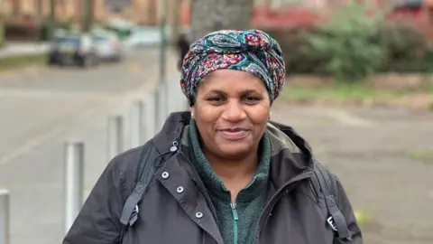 A woman is wearing a turquoise and pink patterned headscarf and silver earrings. She's wearing a zipped green fleece and a grey raincoat. She has a black backpack on.