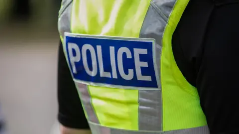 The back of a police officer's vest - it is a yellow high vis with a blue panel reading 'police' across the back, they are wearing a short sleeved black t shirt under the vest and the background is blurred