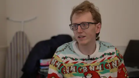 Patrick Hughes/BBC Sam is wearing a bright and colourful jumper and dark-framed glasses. 