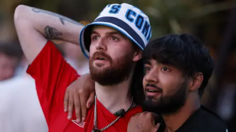 Reuters Two England fans watch the football on a screen, which is not pictured. One is wearing a red England shirt and has his arm rested on his head. His friend has his arms around him.