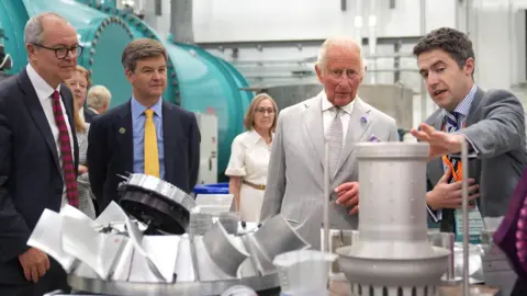 PA Media Several people are standing round different contraptions in a laboratory. King Charles III is among the group and is wearing a pale grey suit and tie while talking to others.