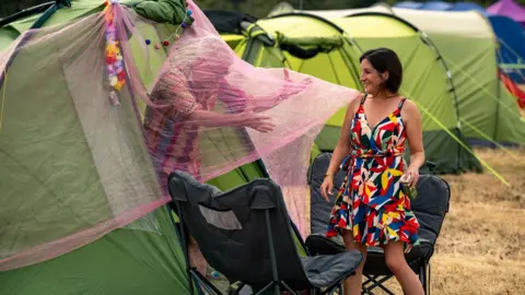 Garry Jones Photography Two campers setting up their green tent, with black camping chairs outside. A man can be seen putting up a large red net across the entrance to the tent, he is reaching forward and laughing. A woman is next to him in a brightly-coloured dress, laughing as he pushes through the material. 