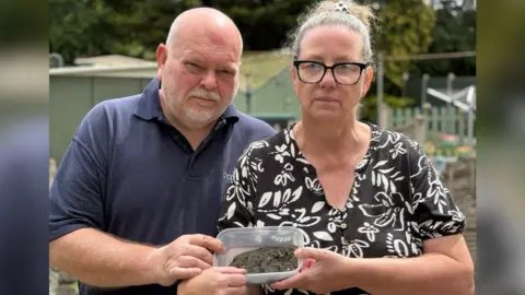 Gary and Debbie Griffin pictured together in a garden. Gary is bald and wearing a blue polo shirt with his collar popped up while Debbie has Thick black framed glasses and her hair is scrapped back in a pony tail. She is wearing a black short sleeved shirt that has white floral prints all over. The couple are holding a plastic clear container with dust in it.
