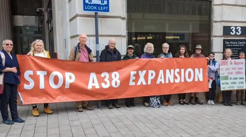 Group of campaigners stood holding an orange banner saying "Stop A38 Expansion"