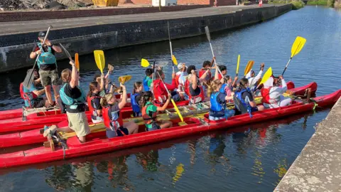 BBC Children in a red bell boat holding up plastic oars.