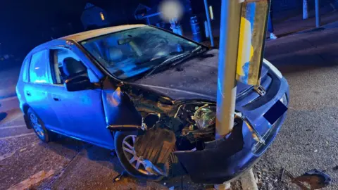 A blue car is badly damaged at the front driver's side. Part of the car is wrapped around a post fitted with traffic lights. There is debris on the road at the crash site.