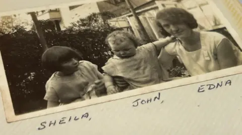 David Turner An old black-and-white photograph of a young girl with short hair in a bob, with a younger boy with light curly hair and a woman with short dark hair. They appear to be sitting in a garden. The picture is labelled: "SHEILA, JOHN, EDNA"