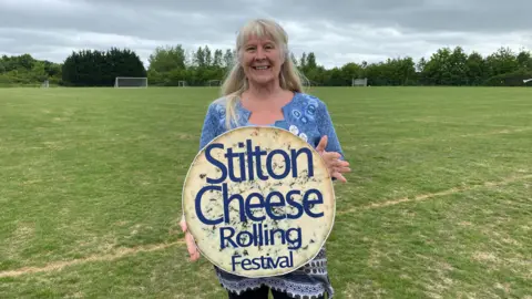 Natalie Malcolm/BBC Tianda Woolner is smiling at the camera standing on some grass and holding a sign saying "Stilton Cheese Rolling Festival". She has long, fair hair and is wearing a blue patterned top.