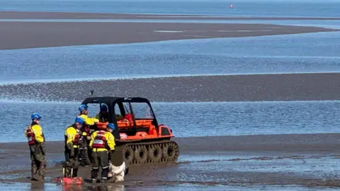 HM Coastguard Fleetwood Three coastguard volunteers in rescue kit extricate the woman from mudflats with tidal channels and the sea in the background.