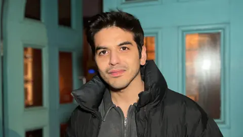 Ahmad is wearing a black padded jacket stands in front of a large turquoise double door. Warm interior light glows through the glass panels behind him.