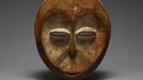 A mask depicting a stylised version of a human face - there is a small smile, narrow eyes and dots pressed into surface which go in a horseshoe shape where eyebrows would be and are also under the nose.