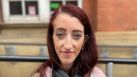 A girl with red hair and facial piercings looking at the camera, with a pink jumper on and head phones on 