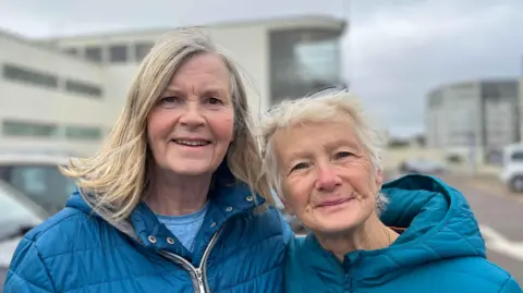 George Carden/BBC Sue and Jane smiling at the camera in the car park with the pavilion entrance behind them. It has a large curved window 