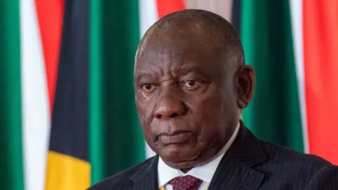 A head and shoulders image of Cyril Ramaphosa wearing a white shirt and red tie, with a pin-striped jacket. Behind him can be seen South African flags