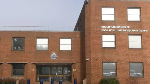 A red brick building with the words Bedfordshire Police Headquarters in white capital letters. An entrance area sits below the force's large emblem which is attached to the wall. 