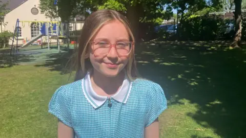 Mikayla ha wearing a light blue and white checked top with a white polo collar. She has long light brown hair and is wearing pink framed glasses.