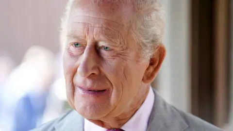 King Charles III wearing a pink shirt and light grey suit. He has receding white hair.