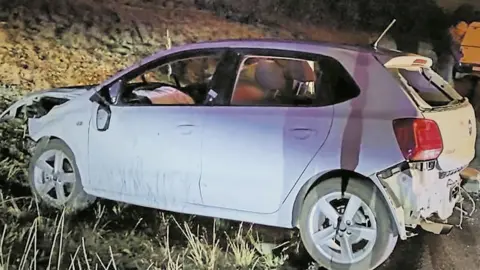 South African police A small white car - damage visible at both ends of the vehicle - on a grass verge